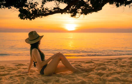 Back view of sexy woman sit on sand and watching sunset at tropical beach. Woman wear swimsuit, sunglasses, and straw hat relaxing at tropical paradise beach on summer vacation. Holiday travel alone.の写真素材