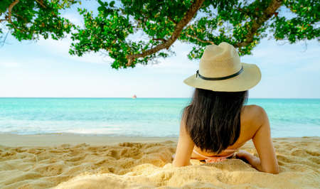Back view of happy young Asian woman in pink swimsuit and straw hat relax and enjoy holiday at tropical sand beach under the tree. Girl in summer vacation fashion. Beauty sexy model. Summer vibes.の写真素材