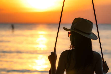 Silhouette woman wear bikini and straw hat swing the swings at the beach on summer vacation at sunset. Girl in swimwear sit on swings and watch beautiful sunset. Summer vibes. Woman travel alone.の写真素材