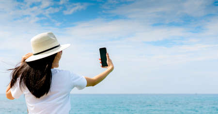 Young Asian woman wear hat in casual style use smartphone taking selfie at pier. Summer vacation at...