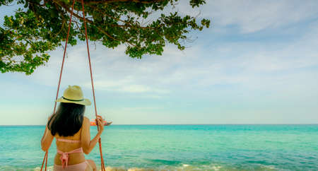 Asian woman wear swimwear and hat swing the swings at sand beach  and looking beautiful tropical paradise sea and sky on sunny day. Summer vacation. Summer vibes. Enjoying and relaxing girl on holidayの写真素材