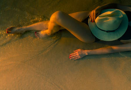 Sexy Asian woman with hat lying down on sand beach during sunset. Woman wear black swimsuit relaxing...