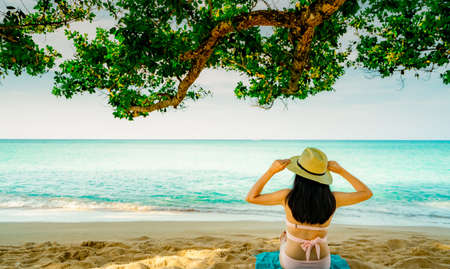 Back view of happy young Asian woman in pink swimsuit and straw hat relax and enjoy holiday at tropical sand beach under the tree. Girl in summer vacation fashion. Beauty sexy model. Summer vibes.の写真素材