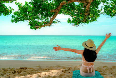 Back view of happy young Asian woman in pink swimsuit and straw hat relax and enjoy holiday at tropical sand beach under the tree. Girl in summer vacation fashion. Beauty sexy model. Summer vibes.の写真素材