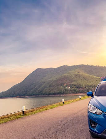 Front view of blue SUV car with opened car truck parked on asphalt road at dam. Summer road trip...