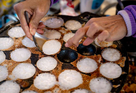 Mortar toasted pastry or Kanom Krok is Thai traditional dessert. Woman hand removing Kanom Krok from stove by spoon. Street food in Thailand. Thai dessert made from coconut milk and flour.の写真素材