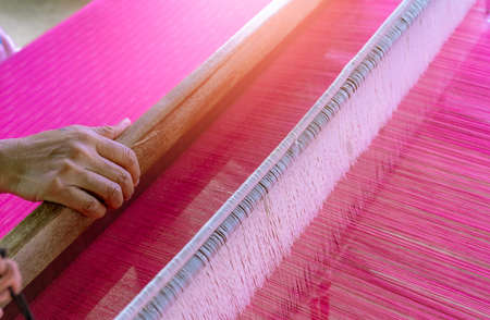Woman working on weaving machine for weave handmade fabric. Textile weaving. Weaving using traditional hand weaving loom on the long cotton strands. Textile production in Thailand. Asian culture.の写真素材