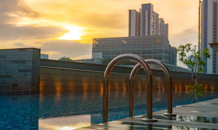 Swimming pool at luxury hotel and spa in the morning with golden sunrise sky. Closeup grab bars ladder with clean water and wooden floor at edge of swimming pool. Leisure at hotel poolside concept.のeditorial素材