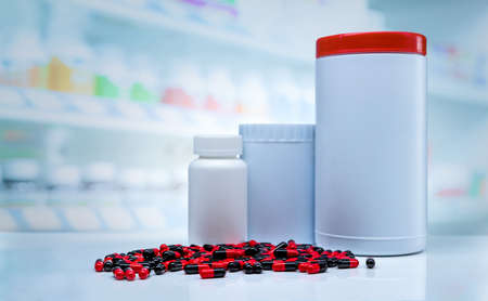 Red-black capsules pills spread on counter in drugstore. Blank label plastic drug bottle on blurred...