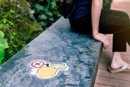 Selective focus on symbol of gap for social distance to prevent coronavirus. Social distancing concept. Blurred woman sit on seat near warning sign for keep distance on public seat at outdoor park.の写真素材