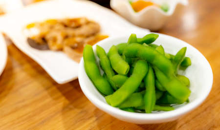 Edamame or green soybeans blanched in white bowl on wooden table at Japanese restaurant. Pods of green soy beans on blurred Japanese food. High fiber and protein food. Plant-based protein. Vegan diet.の写真素材