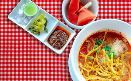 Northern Thai curry noodle soup with chicken and side dish in white bowl on red checkered tablecloth. Top view of Khao Soi and ripe papaya with wooden fork in bowl. Thai traditional food menu.の写真素材