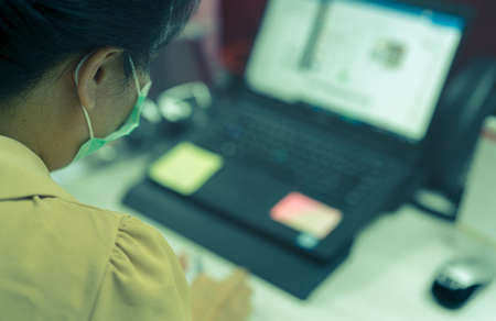 Back view of woman wear face mask working at desk with laptop in office. Asian woman worker using computer at office during work from home. New normal lifestyle in office after coronavirus pandemic.の写真素材