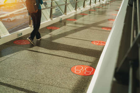 Back view of woman walking on pedestrian glass bridge. Corridor connections between buildings with social distancing symbols on floor for distance footstep. Footsteps sticker stamp on bridge floor.の写真素材