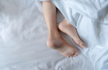 Woman barefoot on bed under white linen blanket in hotel or home bedroom. Healthy sleep and relaxation concept. Lazy Sunday morning. Bare feet of woman chilling sleep on white comfort bed and duvet.の写真素材