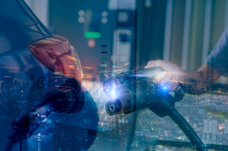 Car charging at electric car charging station and city night lights background. Hand holding EV...