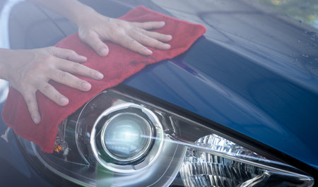 Car wash service. Man hand holding red microfiber cloth and polish headlight of blue car. Auto care service business concept. Man cleaning and detailing luxury car. Clean water drops from luxury car.の写真素材