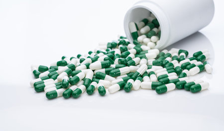 Green and white capsule pills spilled out of a white plastic bottle. Pharmaceutical industry. Prescription drug. Healthcare and medicine. Pharmacy product. Pharmaceutics. Capsule pill manufacture.の写真素材