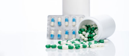 Green-white capsule pills on blur blister pack of blue-white capsule pills and drug bottle. Pills spread out of plastic bottle. Pharmaceutical industry. Prescription drug. Health and medical care.の写真素材