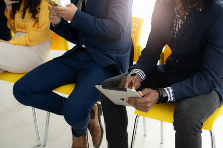 Two men in suits are sitting in chairs, one of them holding a tablet. Concept of professionalism and focus, as the men are likely engaged in a work-related activity. The use of technologyの写真素材