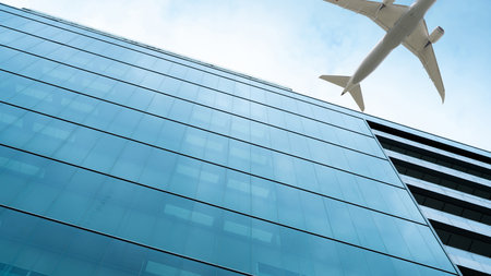 Airplane flying in the sky above modern sustainable glass office building. Exterior skyscraper green glass building. Business travel. Aviation business. Air logistics. Cargo plane. Departure flight.の写真素材