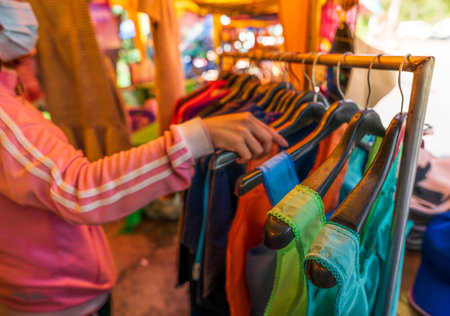 Local Thai market visitor browsing variety of colorful traditional clothing and garments displayed by rural vendors offering authentic handcrafted apparel for tourists and cultural shoppersの写真素材