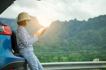 Woman reading book beside blue car with mountain landscape background. Peaceful road trip travel lifestyle and adventure freedom. Solo traveler enjoy reading. Mindful travel and digital detox vacation