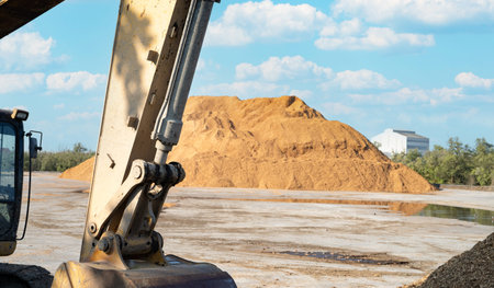 Excavator bucket and hydraulic arm at industrial yard with large sawdust pile, showing heavy...