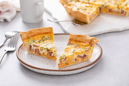 Homemade quiche with ham, cheddar cheese and parsley cut on the table with slices on a plateの写真素材