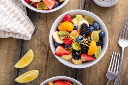 Fruit salad on a wooden table with berries, citrus zest, mint and honey dressing in small bowls overhead viewの写真素材