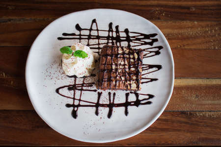 Brownies topped with chocolate and whipped cream. In a white plate on a wooden table, top view.