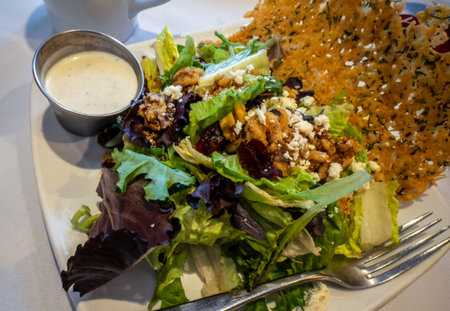 A healthy salad with cheese crisp, dried cranberries, walnuts, feta cheese, and ranch on the white plate with a forkの写真素材