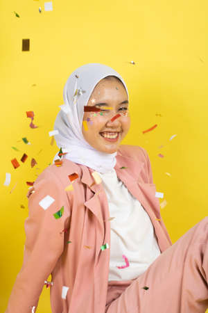 Beautiful young woman throwing confetti and looking happy on yellow background