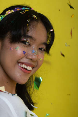 Beautiful young woman throwing confetti and looking happy on yellow background