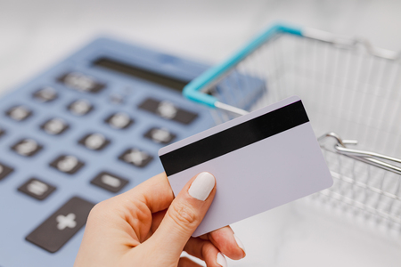 woman's hand holding payment card in front of huge calculator and shopping basket, concept of shopping budgetの写真素材