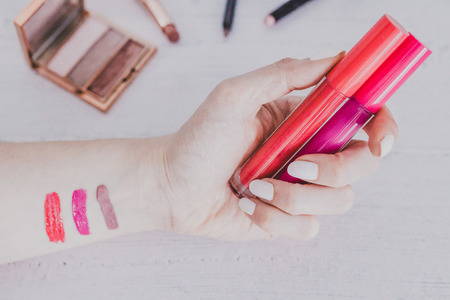 Woman's hand holding lipgloss product and testing it surrounded by other make-up items on desk, concept of beauty industryの写真素材