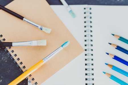 desk with notepads and watercolour brush and coloured pencils with empty pages and copyspace,...