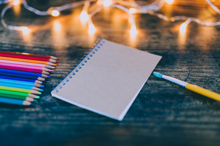 notebook with brushes and color pencils on wooden desk with fairy lights, concept of art and creativityの写真素材