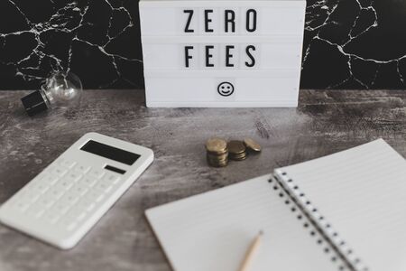zero fees message on lightbox surrounded by coins calculator on office stationery on concrete desk, muted tonesの写真素材