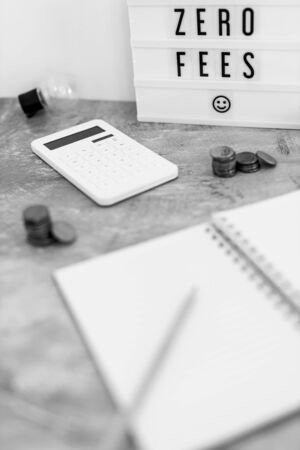 zero fees message on lightbox surrounded by coins calculator on office stationery on concrete desk, black and whiteの写真素材