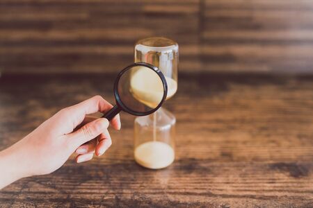 hand holding magnifying glass analyzing time passying by with an hourglass, concept of time management and staying on scheduleの写真素材