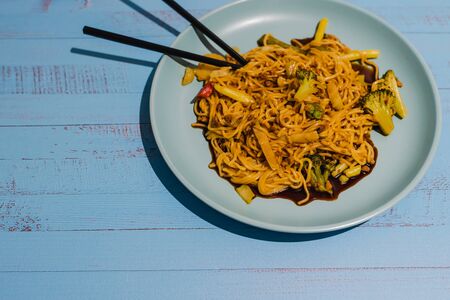 healthy vegetarian noodles with mixed veggies and spicy curry sauce served on teal plate with chopsticks on blue wooden backgroundの写真素材