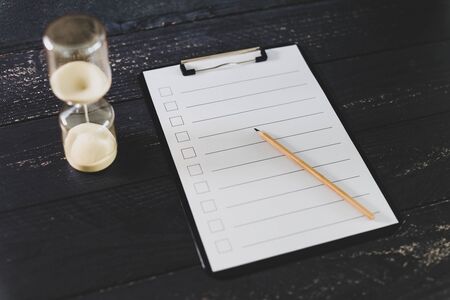 clipboard with blank list with cases to tick off on wooden desk with hourglass, concept of time...