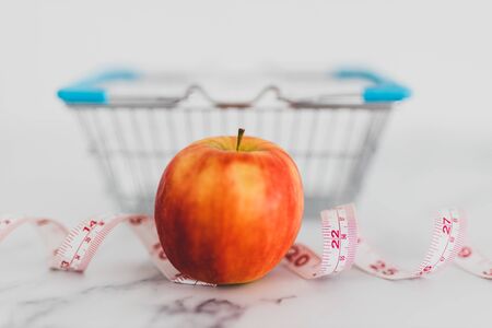 diet and nutrition concept, red apple with measuring tape and grocery store shopping basket shot at shallow depth of fieldの写真素材