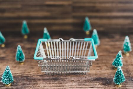 Shopping basket surrounded by miniature Christmas trees, concept of festive season shoppingの写真素材
