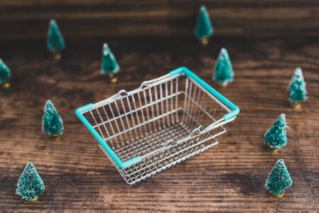 Shopping basket surrounded by miniature Christmas trees, concept of festive season shoppingの写真素材