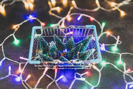 Shopping basket full of miniature Christmas trees and fairy lights all around it, concept of festive season and buying decorationsの写真素材