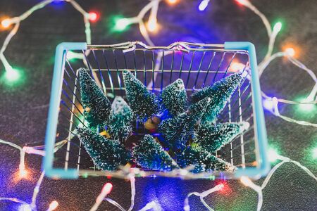 Shopping basket full of miniature Christmas trees and fairy lights all around it, concept of festive season and buying decorationsの写真素材