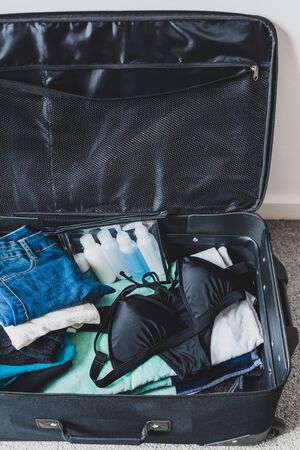 travel essentials and packing for a holiday concept, luggage with pile of clothes beach towel and...