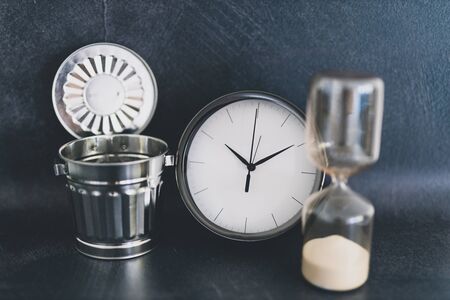 not wasting time conceptual still-life, oversize clock with miniature garbage bin and hourglass on business deskの写真素材